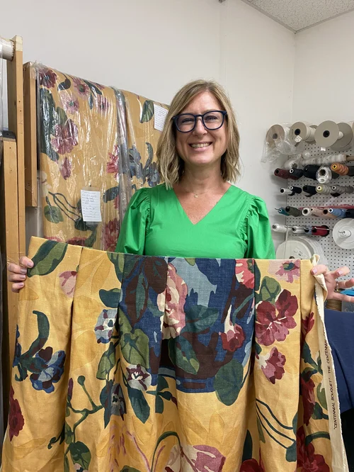 A woman in a green shirt holds up yellow floral fabric in a workshop with fabric rolls and tools visible on shelves behind her, showcasing her expertise in creating custom window treatments for homes in Savannah.