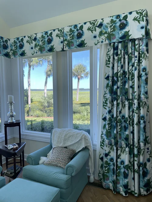 Blue armchair with a white blanket is beside floral curtains and large windows with custom window treatments, overlooking palm trees and greenery.