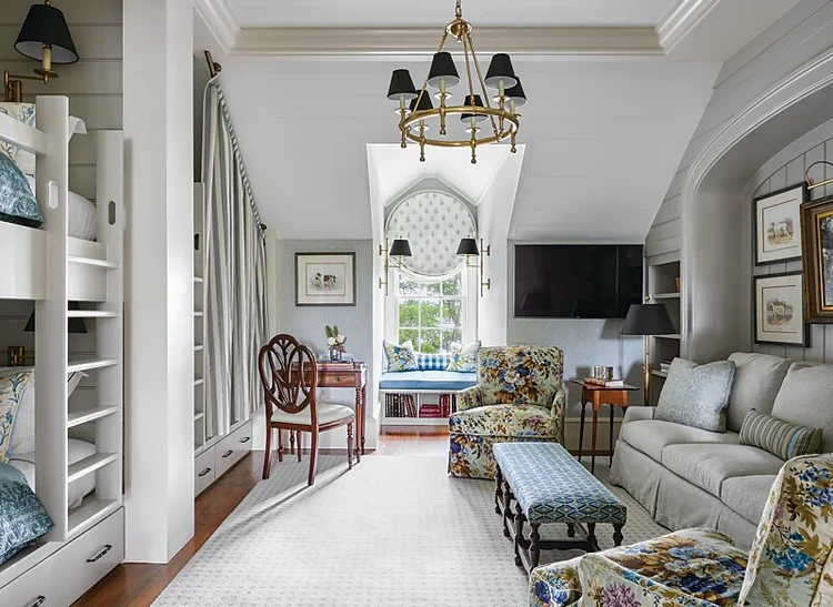 A cozy living area with a sofa, floral armchairs, bunk beds, a desk by a window adorned with elegant custom window treatments, bookshelves, and a chandelier, all in a softly lit attic room with neutral and pastel tones.