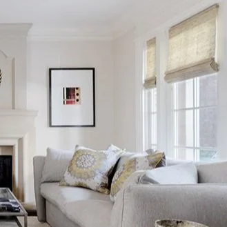 A light gray sofa with patterned cushions sits in a bright living room, next to windows featuring elegant custom window treatments and a framed abstract artwork on the wall.
