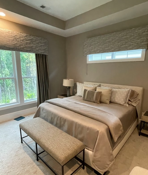 Neutral-colored bedroom with a neatly made bed, multiple pillows, a bench at the foot, two bedside tables with lamps, and large windows featuring custom window treatments with elegant valances that let in natural light.