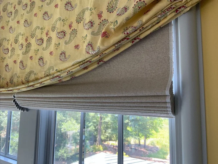 A close-up of a window with a partially raised Roman shade featuring a floral patterned fabric and a beige backing, this elegant design adds charm to your soft furnishings while revealing trees and sunlight outside.