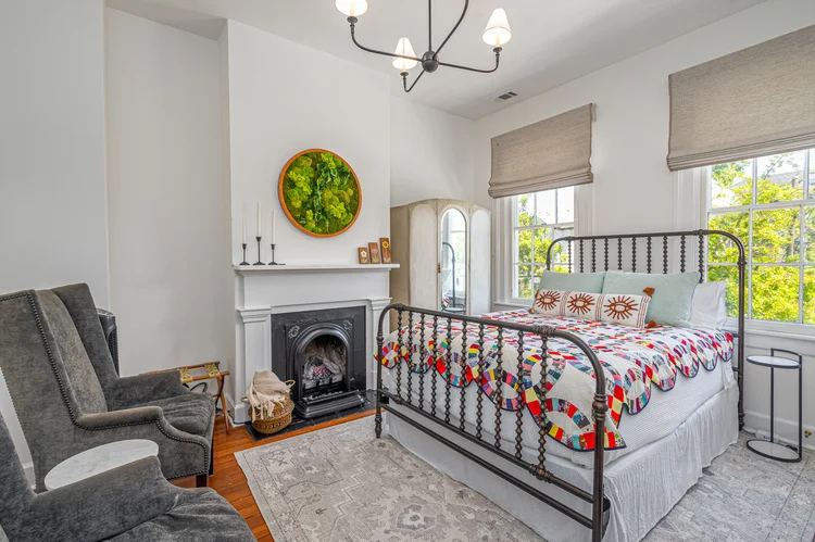 Bright bedroom with a metal bed frame, colorful quilt, fireplace, two gray armchairs, large windows adorned with elegant window treatments, and neutral decor. Soft furnishings throughout the space add comfort and style, creating a cozy retreat inspired by the charm of Savannah.