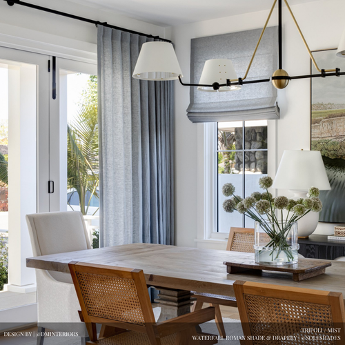 Modern dining area with a wooden table, woven-back chairs, gray drapes, a Roman shade from our custom window treatments collection, a contemporary chandelier, and a glass vase with white flowers on the table—bringing elegant soft furnishings to your Savannah home.