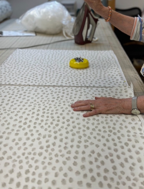 Person smoothing white fabric with gray dots on a worktable, thoughtfully working on a new design for soft furnishings, with an iron, yellow pincushion, and sewing tools visible nearby.