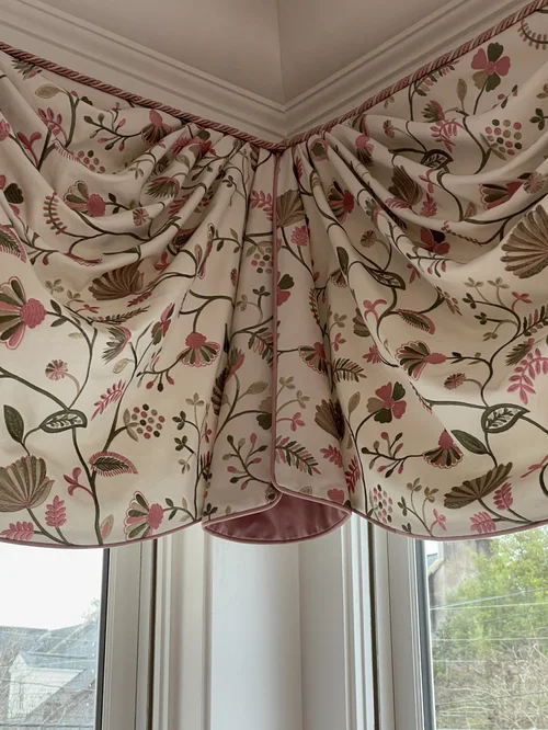 Floral-patterned, scalloped valance drapes hang above corner windows as part of the custom window treatments, featuring pink, green, and beige tones with outdoor scenery partially visible below.