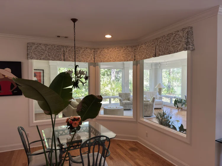 Dining area with a glass table and chairs next to large interior windows featuring elegant window treatments, overlooking a bright sunroom with plants, seating, and thoughtfully coordinated soft furnishings.