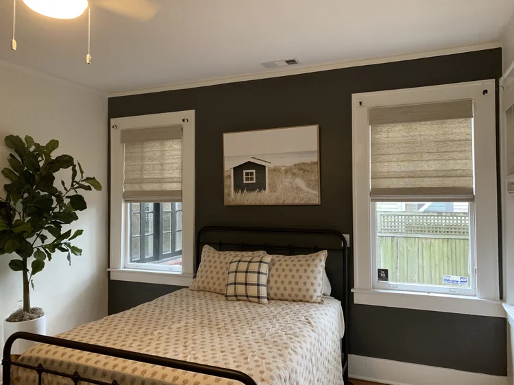 A neatly made bed with patterned bedding sits between two windows dressed in elegant custom window treatments with beige shades. A potted plant is on the left, and a photo of a small house hangs on the dark accent wall, adding a touch of Savannah charm.