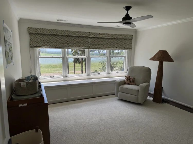 A bright room with a large window featuring elegant window treatments, white walls, a beige recliner, a tall floor lamp, and a wooden cabinet. The thoughtful design allows the window to overlook a grassy savannah and water outside.