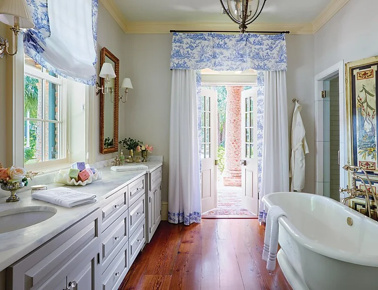 Bright bathroom with double sink, marble countertops, white cabinets, freestanding bathtub, wooden floor, and doors open to a garden. Blue and white valances serve as stylish window treatments for the windows and door, enhancing the room’s elegant design.
