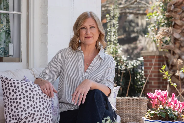 A woman with shoulder-length blond hair sits on a porch sofa with patterned pillows and soft furnishings, wearing a striped shirt and dark pants. Pink flowers and greenery are visible in the background, creating a relaxing savannah-inspired setting.