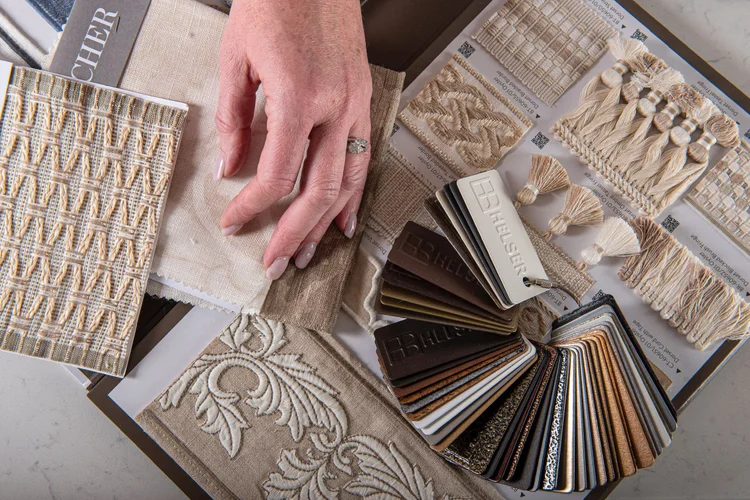 A hand touches fabric swatches next to trim samples and a color fan deck, showing various neutral textile and decorative options for custom window treatments and soft furnishings in elegant design schemes.