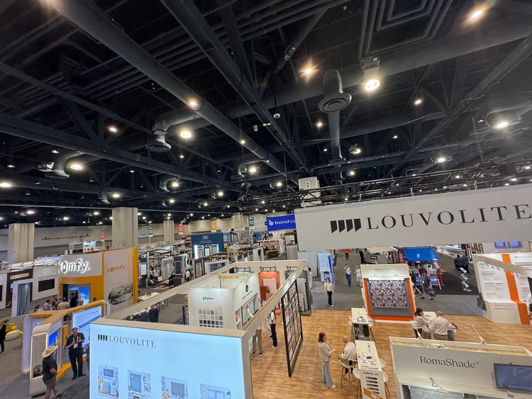 A large indoor trade show with multiple booths and displays showcases the latest in design, including innovative soft furnishings and stylish window treatments, as attendees walk around under bright overhead lighting that illuminates the convention space.