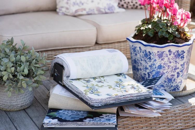 Fabric swatch books and samples on a wooden table with two potted plants, in front of a beige outdoor sofa with plush cushions—perfect inspiration for your next window treatments design, evoking the serene tones of the savannah.