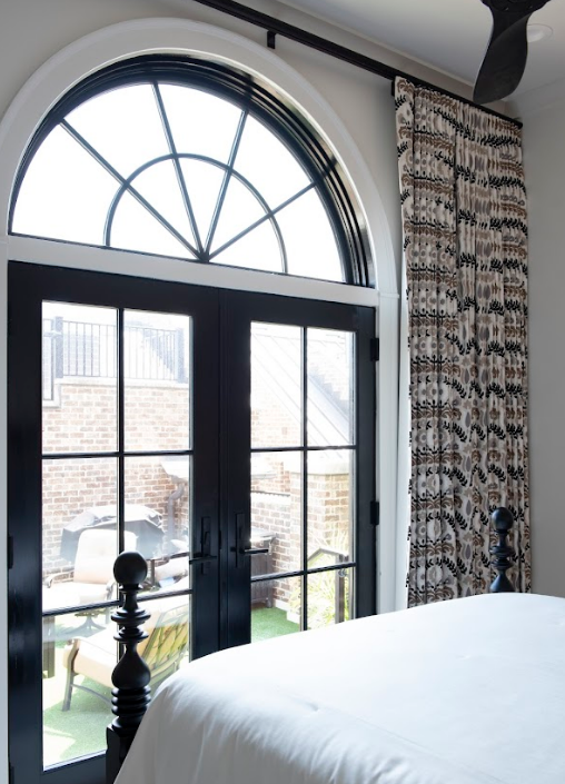 Arched black-framed glass doors with a fanlight window open to a brick patio, complemented by patterned curtains and custom window treatments in this bright, design-forward bedroom.