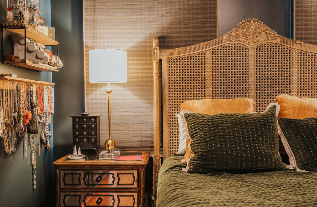 A decorated bedroom with a carved headboard, olive green bedding, soft furnishings that add comfort and style, a wooden nightstand with a lamp, and jewelry displayed on shelves and hooks on the wall. Custom window treatments complete the look by enhancing both privacy and elegance.