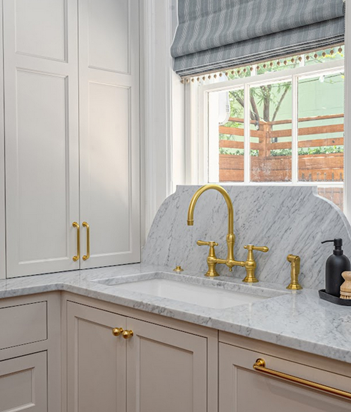 A white kitchen sink with a marble countertop and backsplash, brass faucet and handles, light cabinets, and a window above the sink adorned with custom window treatments in a striped fabric shade creates a cohesive design enhanced by thoughtfully chosen soft furnishings.