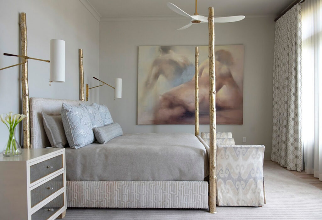 Modern bedroom with a four-poster bed, wall-mounted lamps, abstract art above the headboard, and luxurious soft furnishings that enhance the neutral bedding. A nightstand with flowers sits beside a patterned armchair near a large window adorned with custom window treatments and elegant curtains.