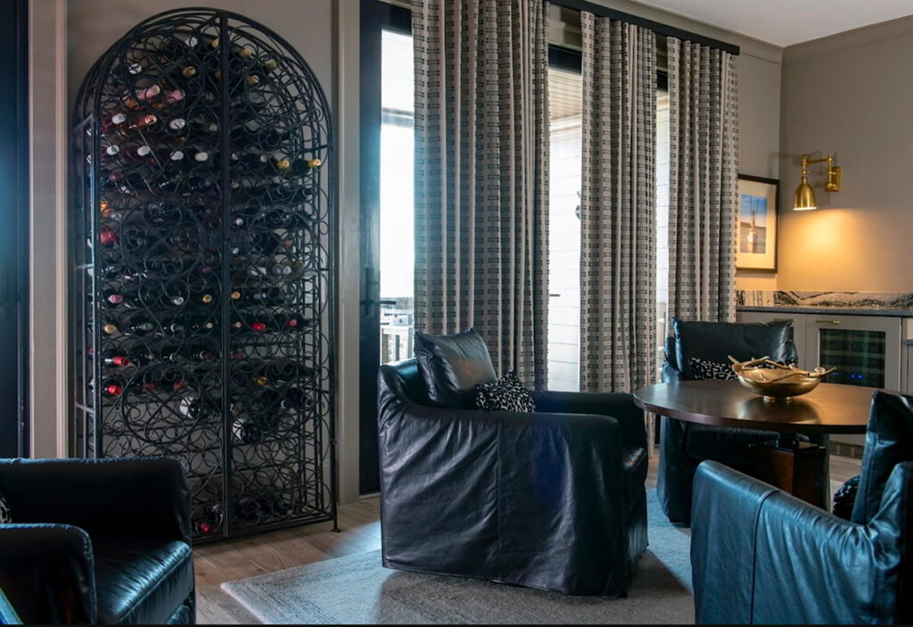 A modern living room features black leather chairs, a round table, a tall wine rack filled with bottles, large windows with patterned window treatments, and a wall-mounted light fixture—all coming together in a cohesive design.