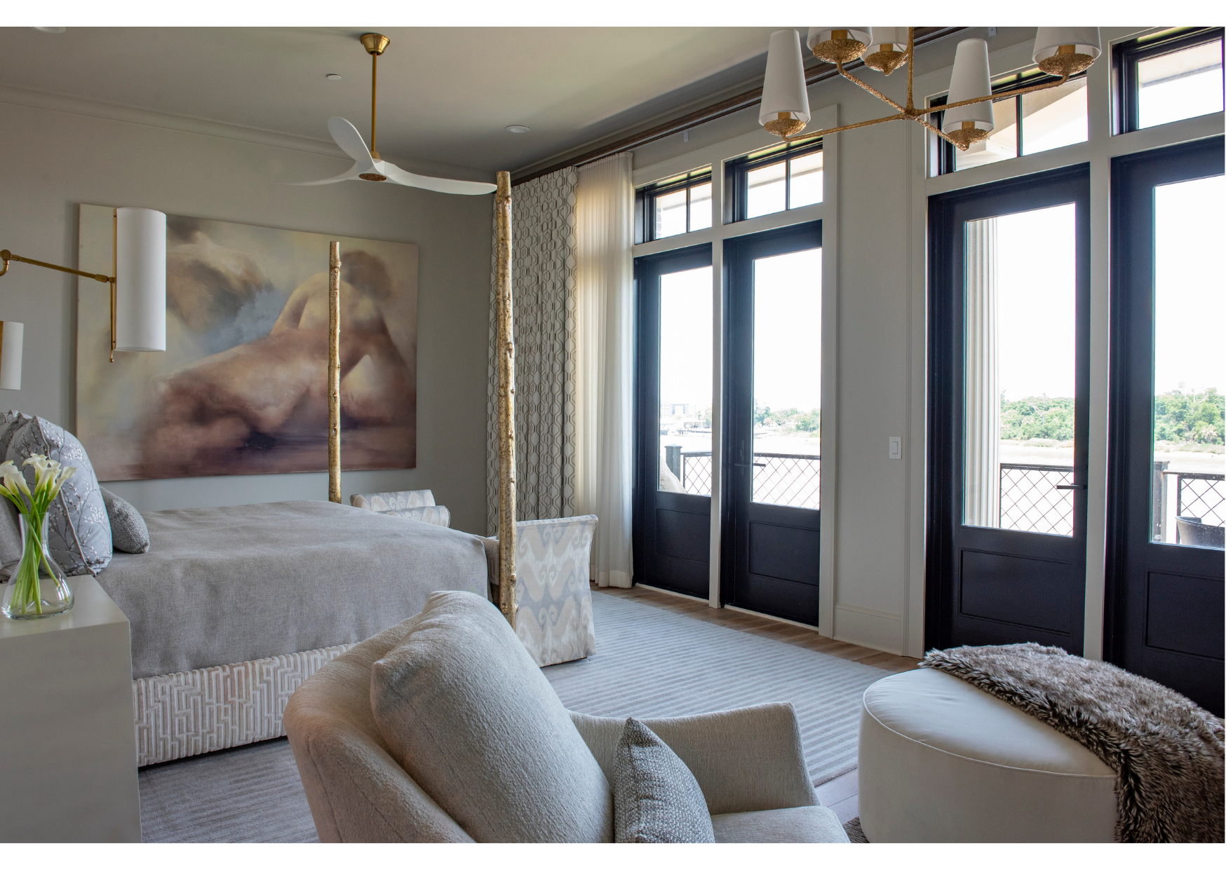 Modern bedroom with large windows and doors adorned with custom window treatments, neutral-toned furniture, gold accents, a ceiling fan, abstract wall art, and a view of greenery outside.