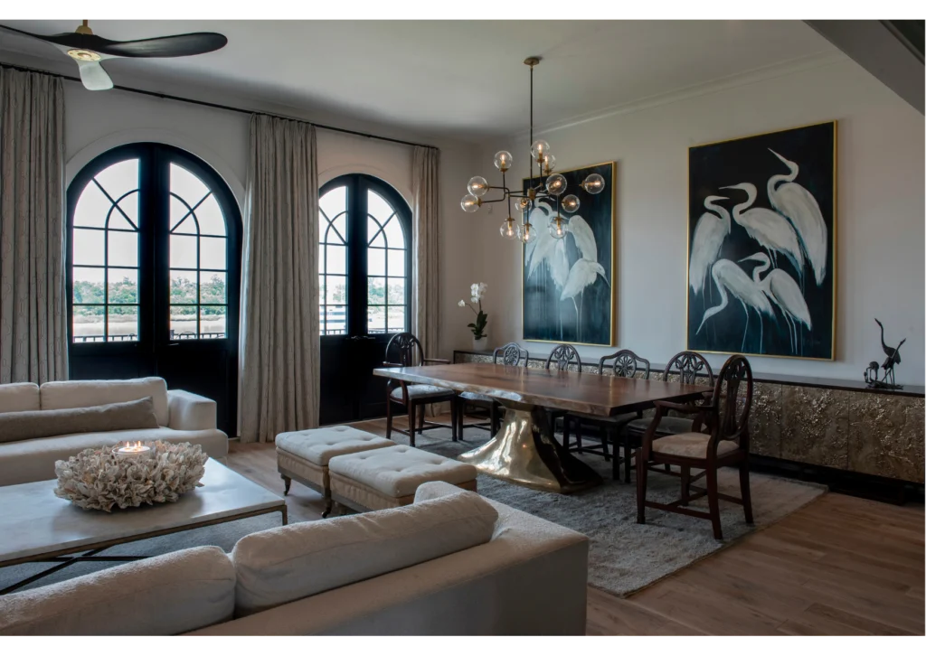 Modern dining and living area with arched windows, beige sofas adorned with elegant soft furnishings, a long wooden table, gold accents, stylish window treatments, and two large white heron paintings on the wall.