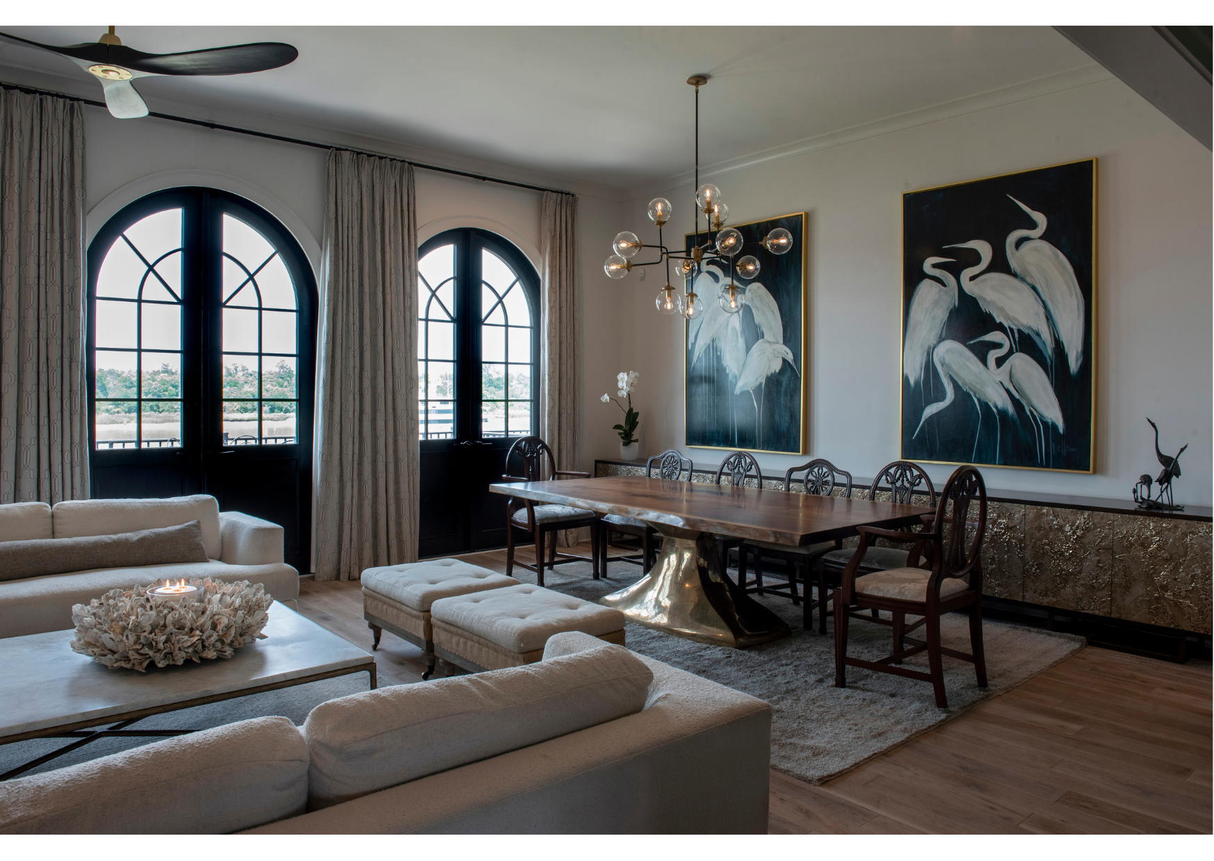 Modern dining and living area with arched windows, beige sofas adorned with elegant soft furnishings, a long wooden table, gold accents, stylish window treatments, and two large white heron paintings on the wall.