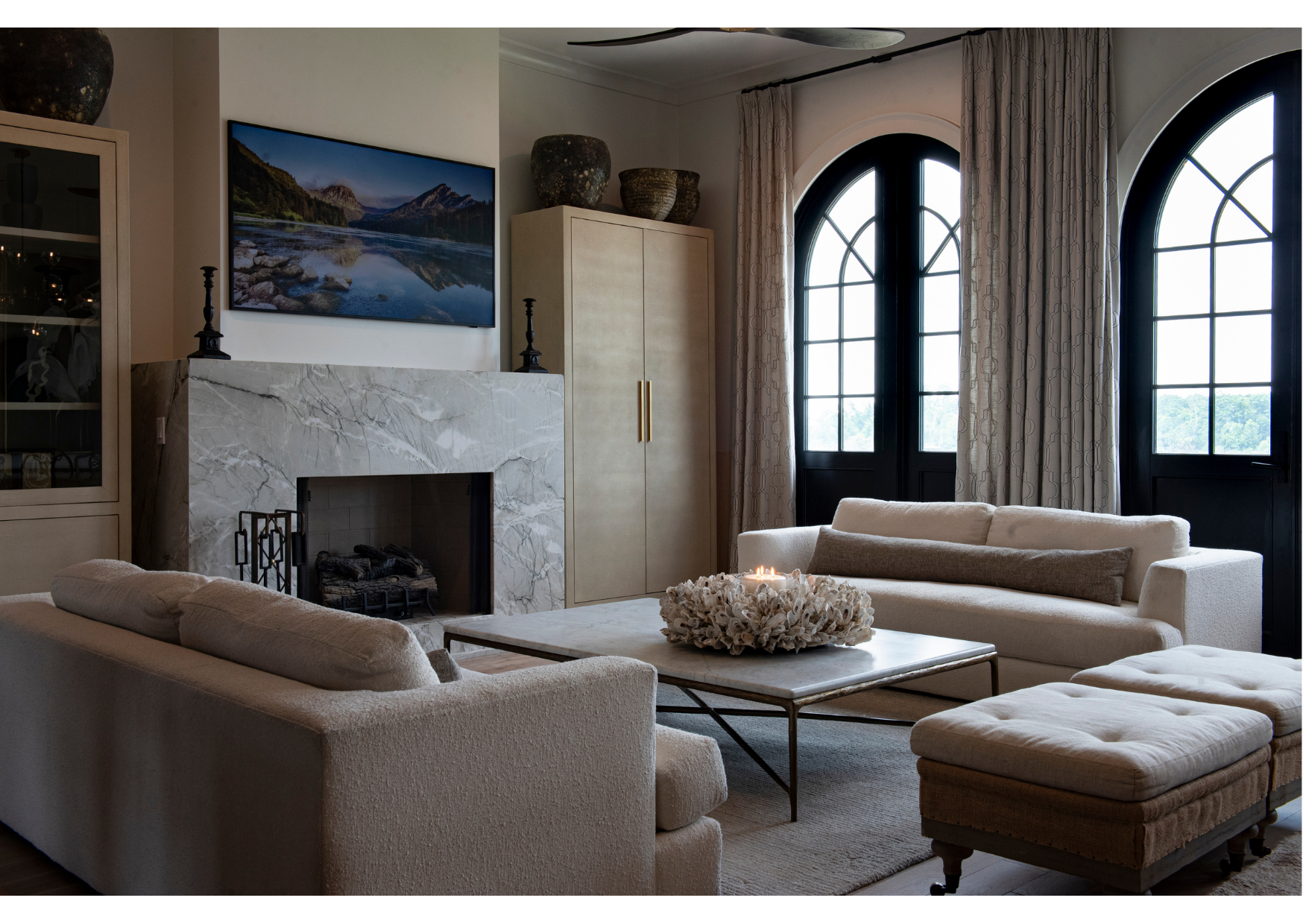 Modern living room with two beige sofas, a marble fireplace, a wall-mounted TV, a square coffee table, and large arched windows with elegant window treatments and coordinating soft furnishings.