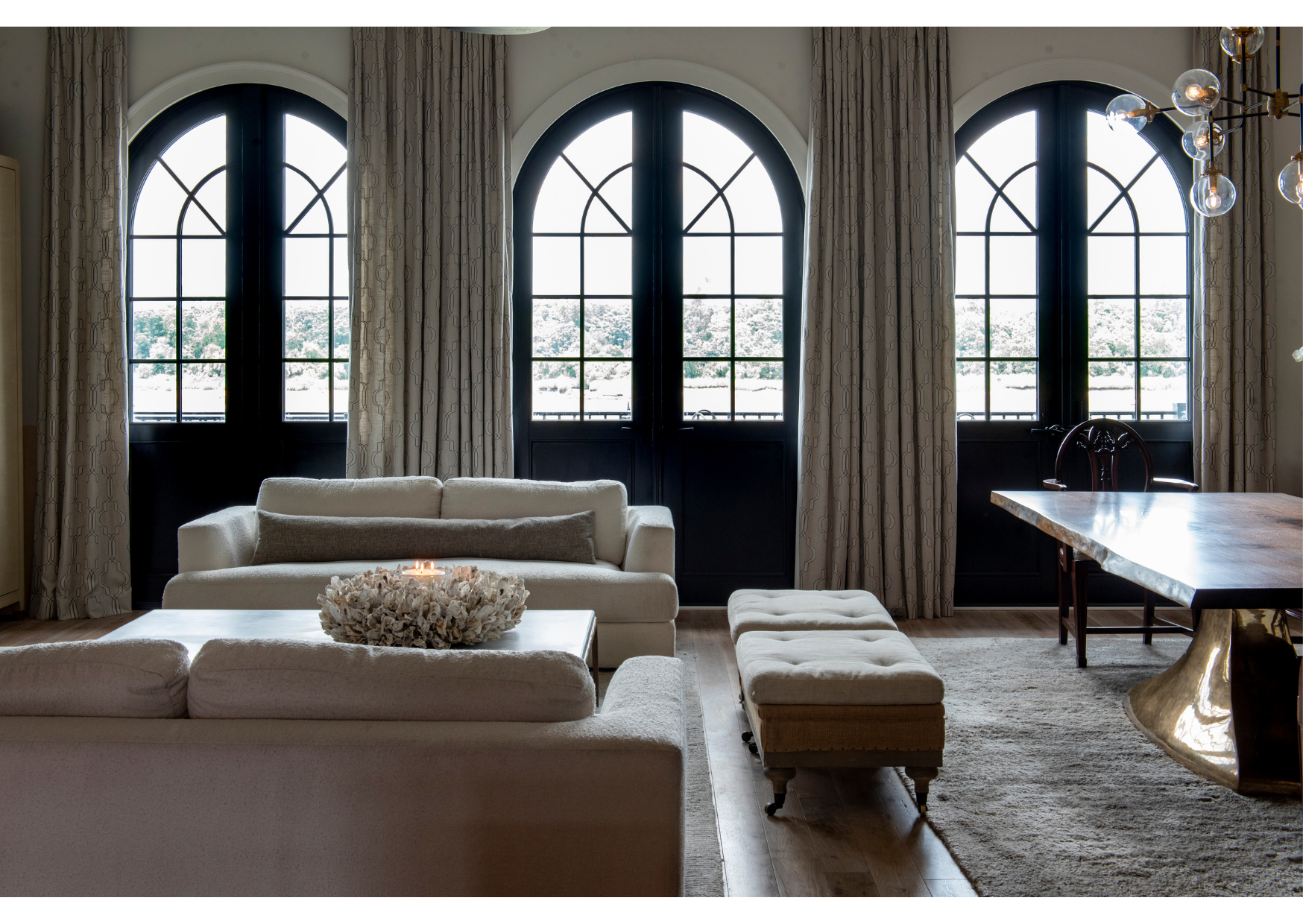 Modern living room with two white sofas, an ottoman, a wood dining table, arched windows with elegant window treatments, and a decorative centerpiece on the coffee table—all elements that showcase thoughtful design.