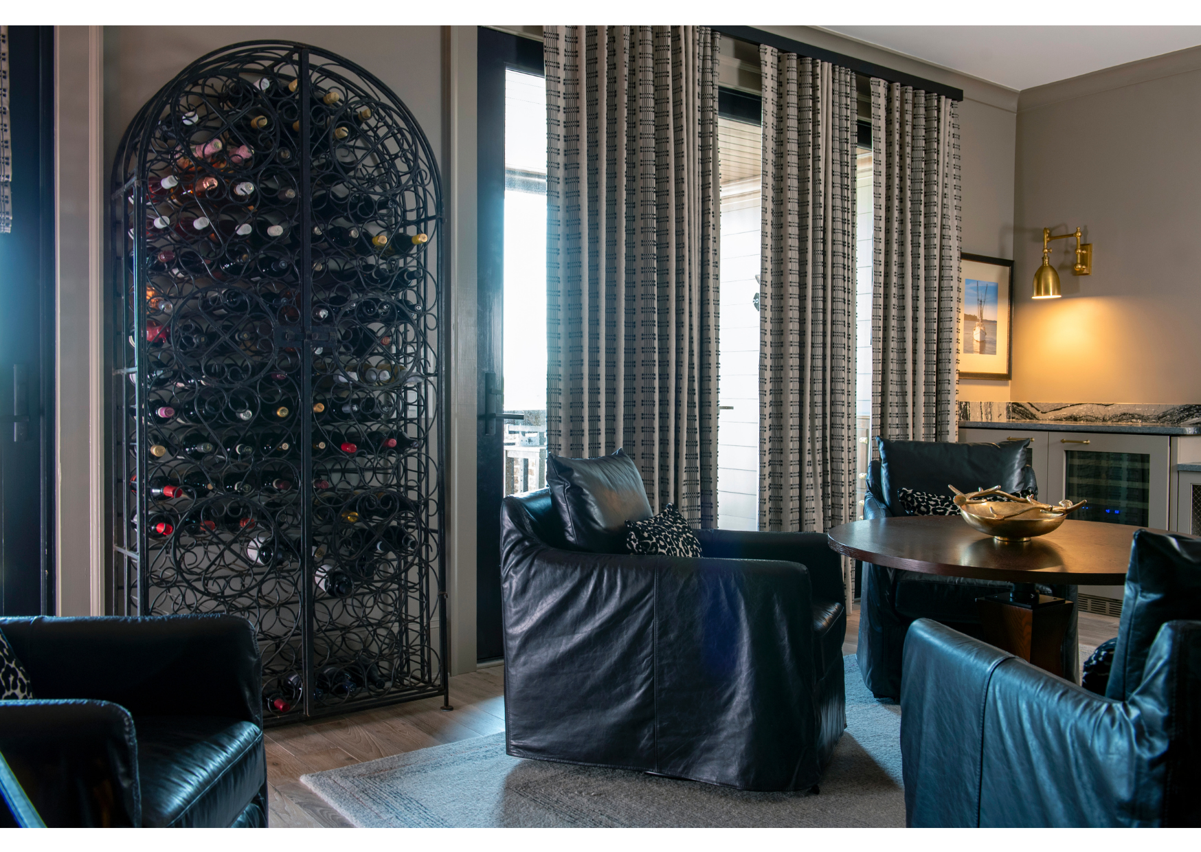 A modern lounge with black leather chairs, a round table, large ornate wine rack, custom window treatments featuring patterned curtains, and a wall-mounted light fixture.