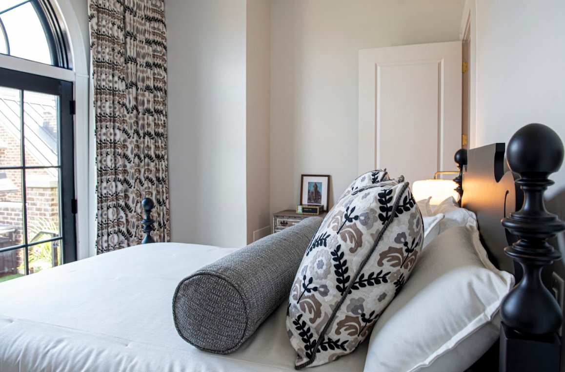 A neatly made bed with patterned pillows, a cylindrical cushion, a side table with a framed photo, and custom window treatments in the form of patterned curtains beside an arched window in a bright bedroom.