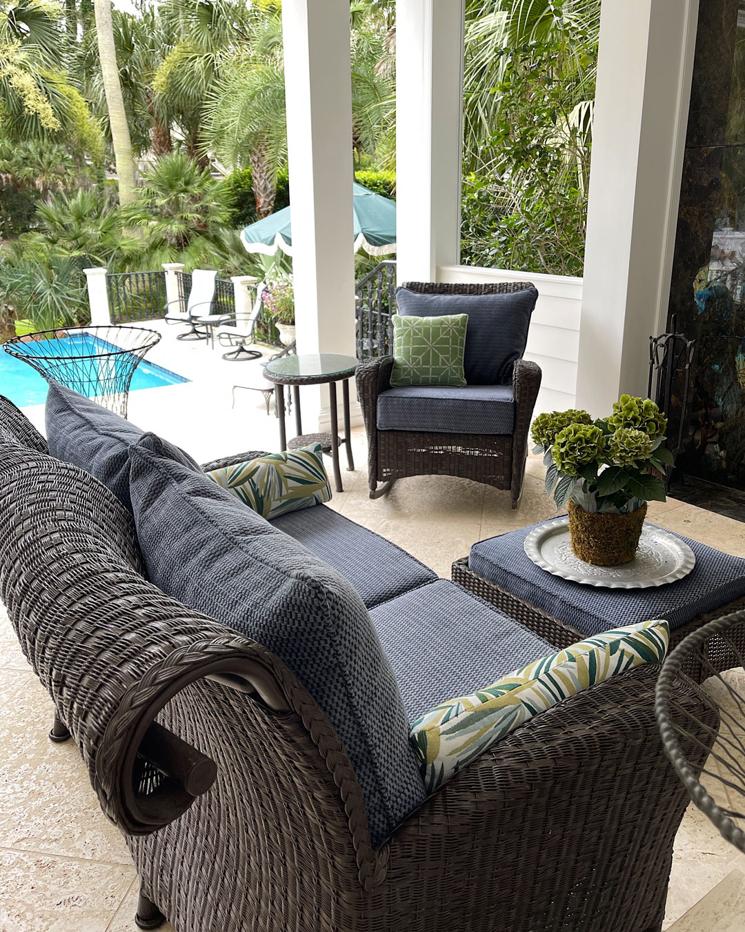 Wicker patio furniture with blue cushions and patterned pillows on a covered terrace, complemented by elegant window treatments, with a pool and greenery visible in the background.