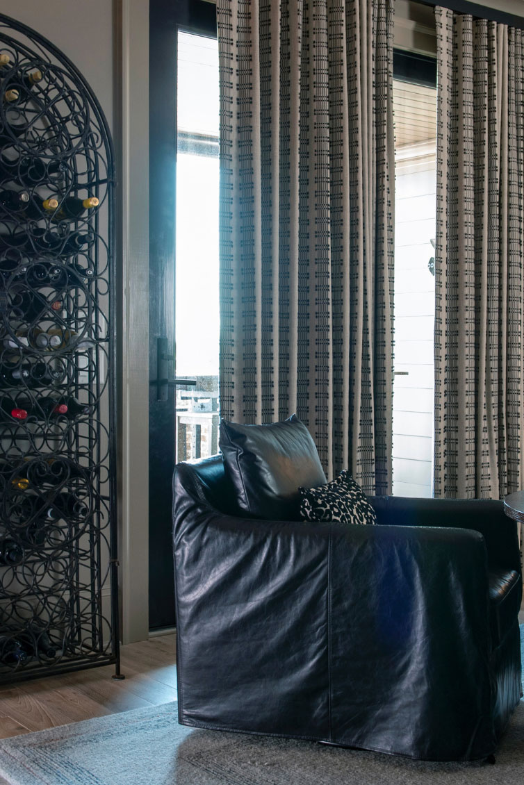 Black leather chair next to patterned curtains and a tall metal wine rack holding multiple bottles in a modern living room accented with soft furnishings for added comfort.
