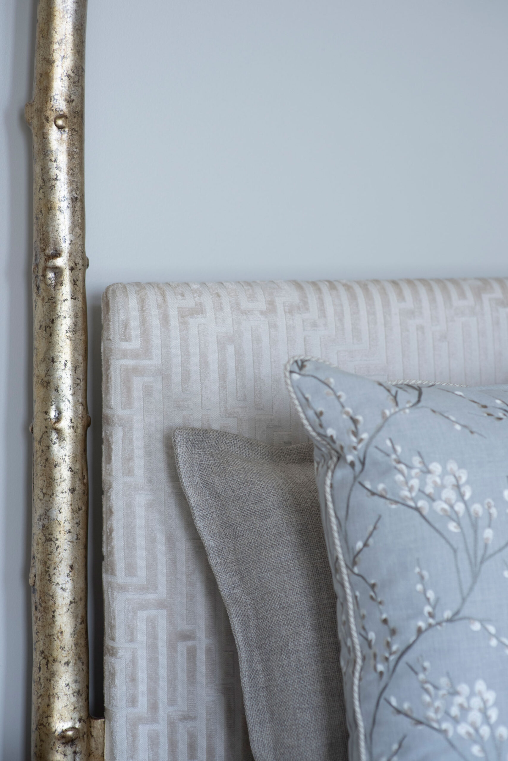 Close-up of a bed with a patterned beige headboard, a gold metallic bedpost, and two pillows—one grey, one light blue with white floral embroidery—showcasing thoughtful design details often found in custom window treatments favored in Savannah homes.