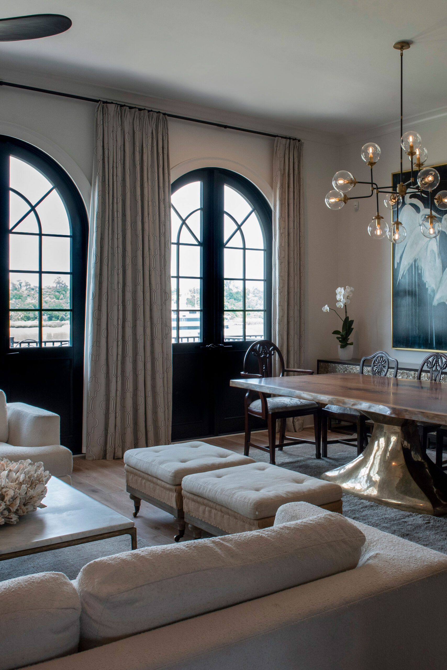 Modern living and dining room with arched windows, light sofas adorned with elegant soft furnishings, a wooden dining table with chairs, a gold base, and a contemporary chandelier. Custom window treatments complement the neutral color palette and flood the space with natural light, showcasing thoughtful design.