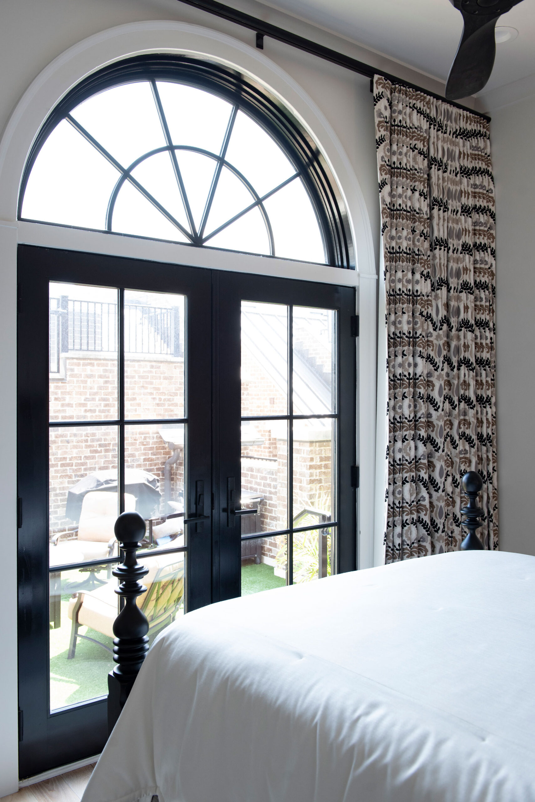Bedroom with a white bed, custom window treatments in the form of patterned curtains, and large black French doors with an arched window above, opening to a brick patio with outdoor furniture.