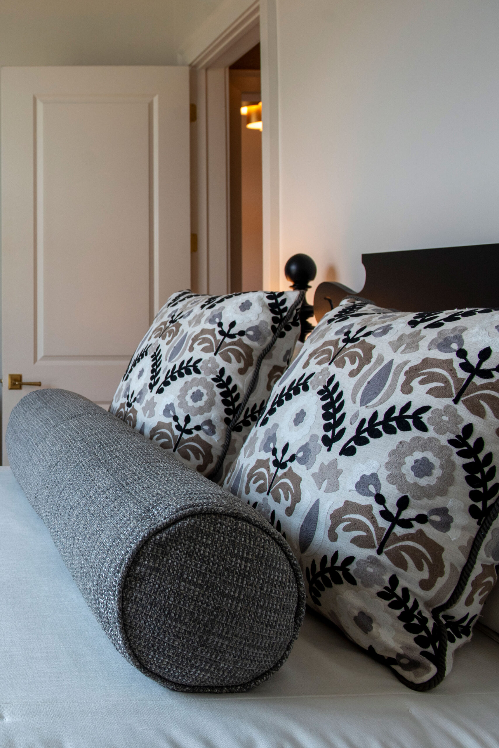 A close-up of a bed with two floral-patterned pillows and a cylindrical gray bolster pillow, complemented by elegant soft furnishings, positioned next to an open white door.