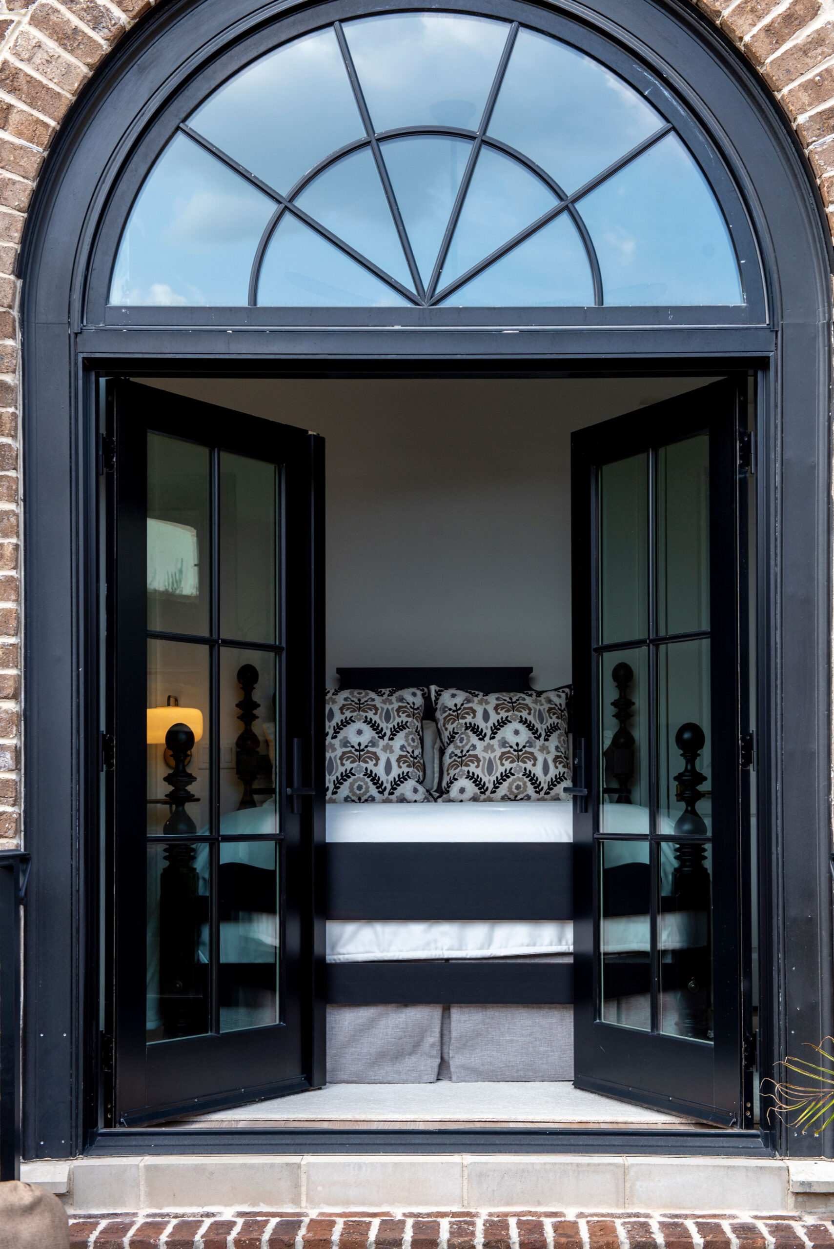 A bed with patterned pillows and luxurious soft furnishings is visible through open black French doors beneath a large arched window with a brick frame.