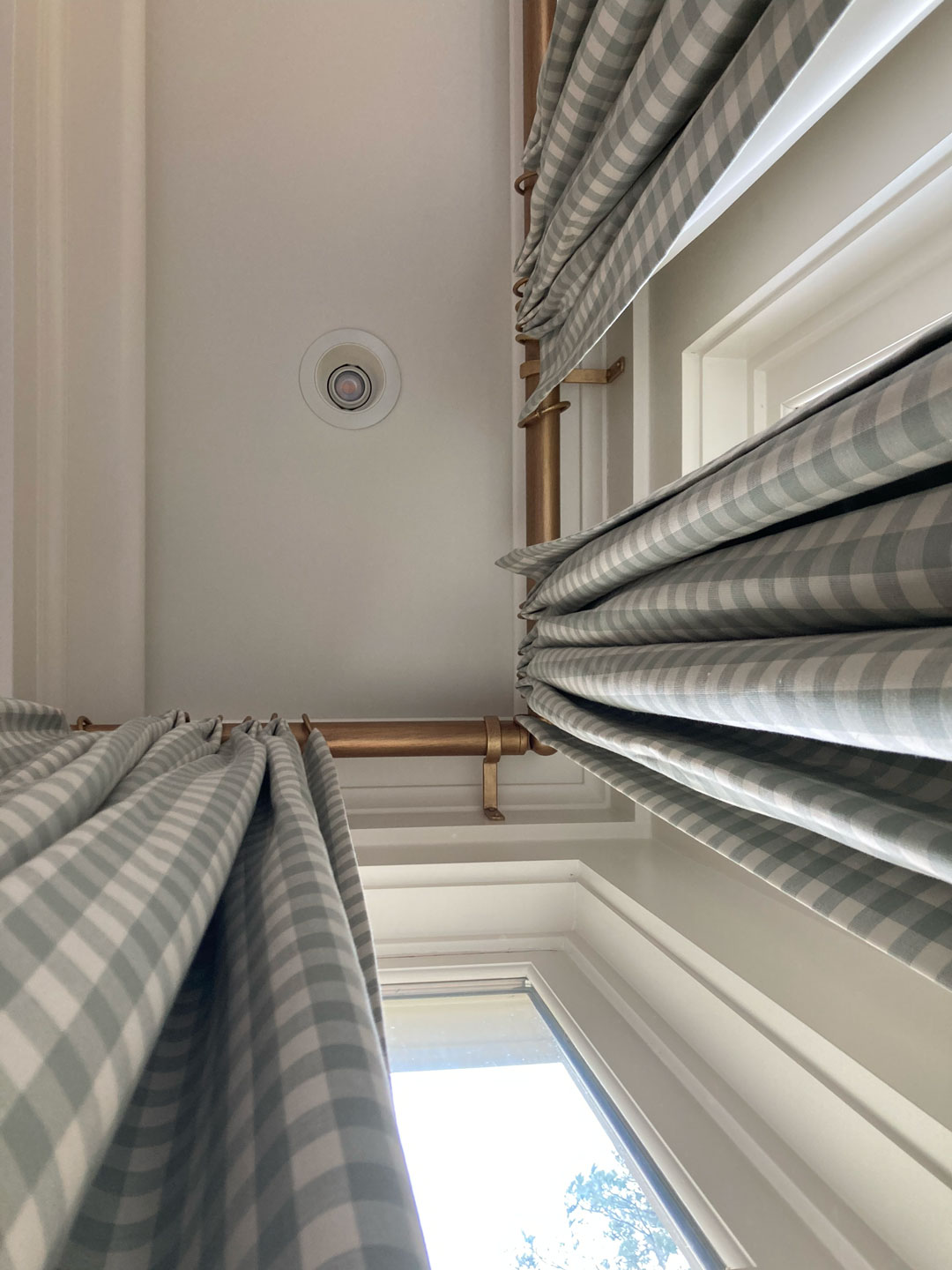 View looking up at ceiling, smoke detector, gold curtain rods, and blue gingham curtains—custom window treatments that bring a touch of savannah charm—with a window partially visible at the bottom.