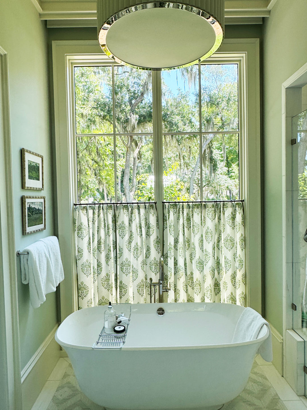 Freestanding bathtub beneath large window with leafy window treatments, small table with bath items, towel on rack, and framed pictures on pale green wall. Trees are visible outside.