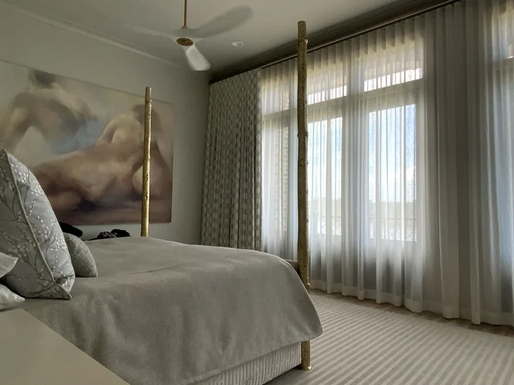 A bedroom with a four-poster bed, neutral bedding, sheer custom window treatments covering tall windows, and a large abstract art piece on the wall.