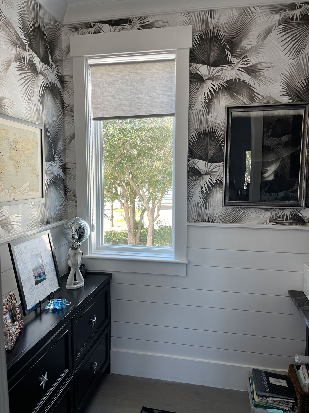 Small room with palm-patterned wallpaper, a window showing trees outside enhanced by elegant window treatments, black furniture, framed photos, a lamp, and various decorative items on the dresser create a thoughtful design inspired by the relaxed charm of a savannah retreat.