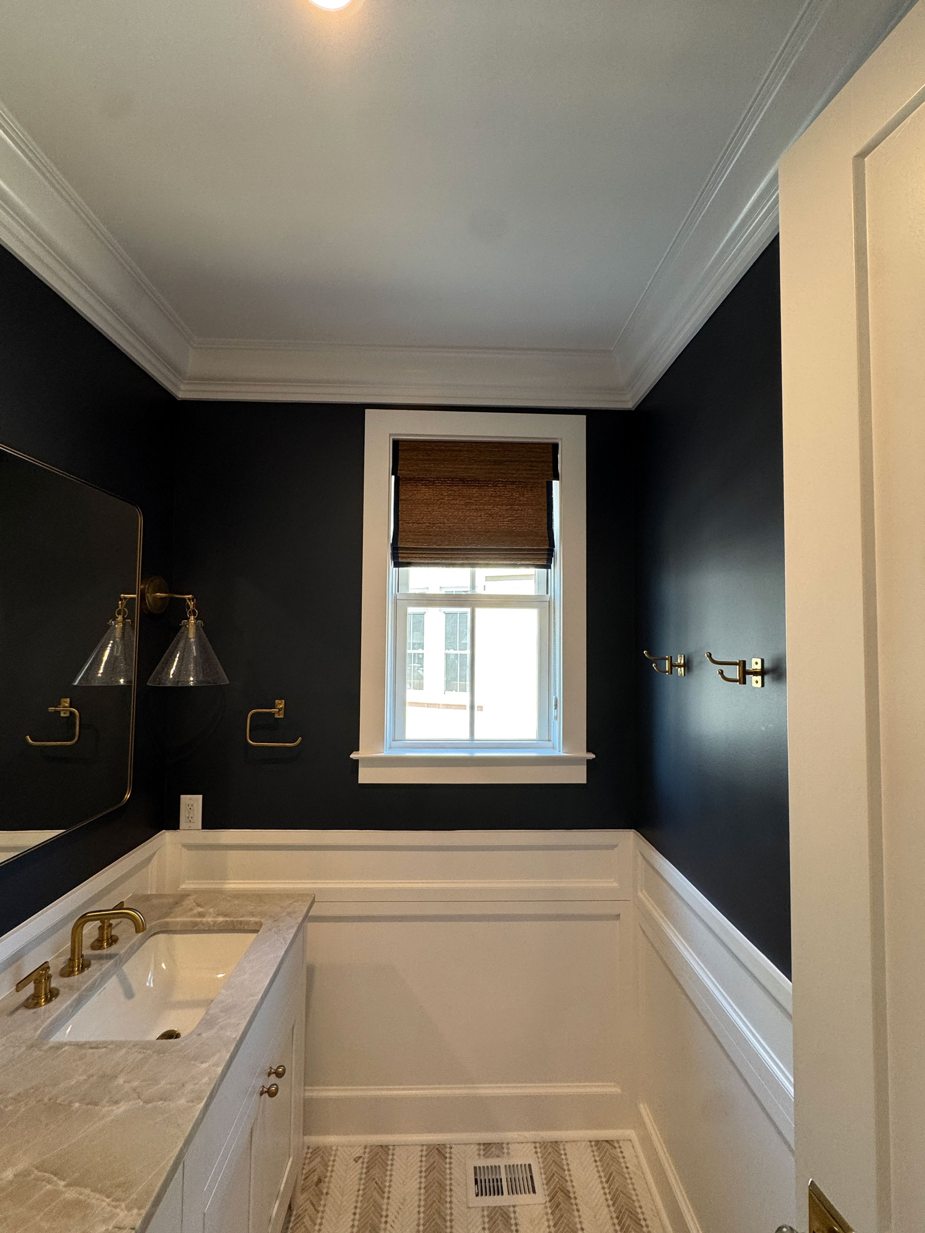 A small bathroom with dark upper walls, white trim, a marble sink, brass fixtures, a window adorned with custom window treatments in the form of a woven shade, and patterned tile flooring highlights thoughtful design throughout the space.