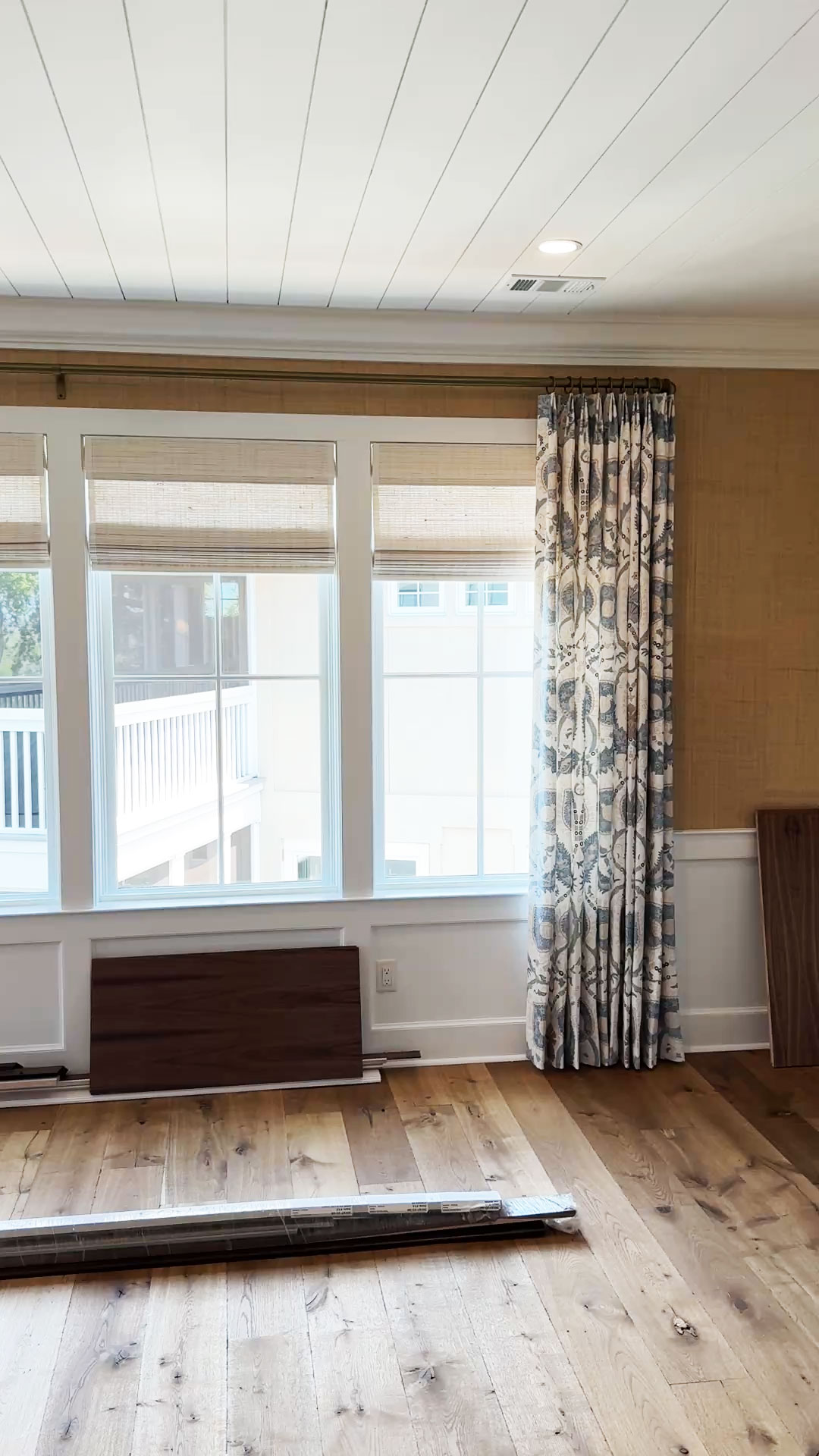 Bright room with large windows, custom window treatments featuring patterned curtains, wood flooring, and wooden panels leaning against the wall, suggesting ongoing renovations.