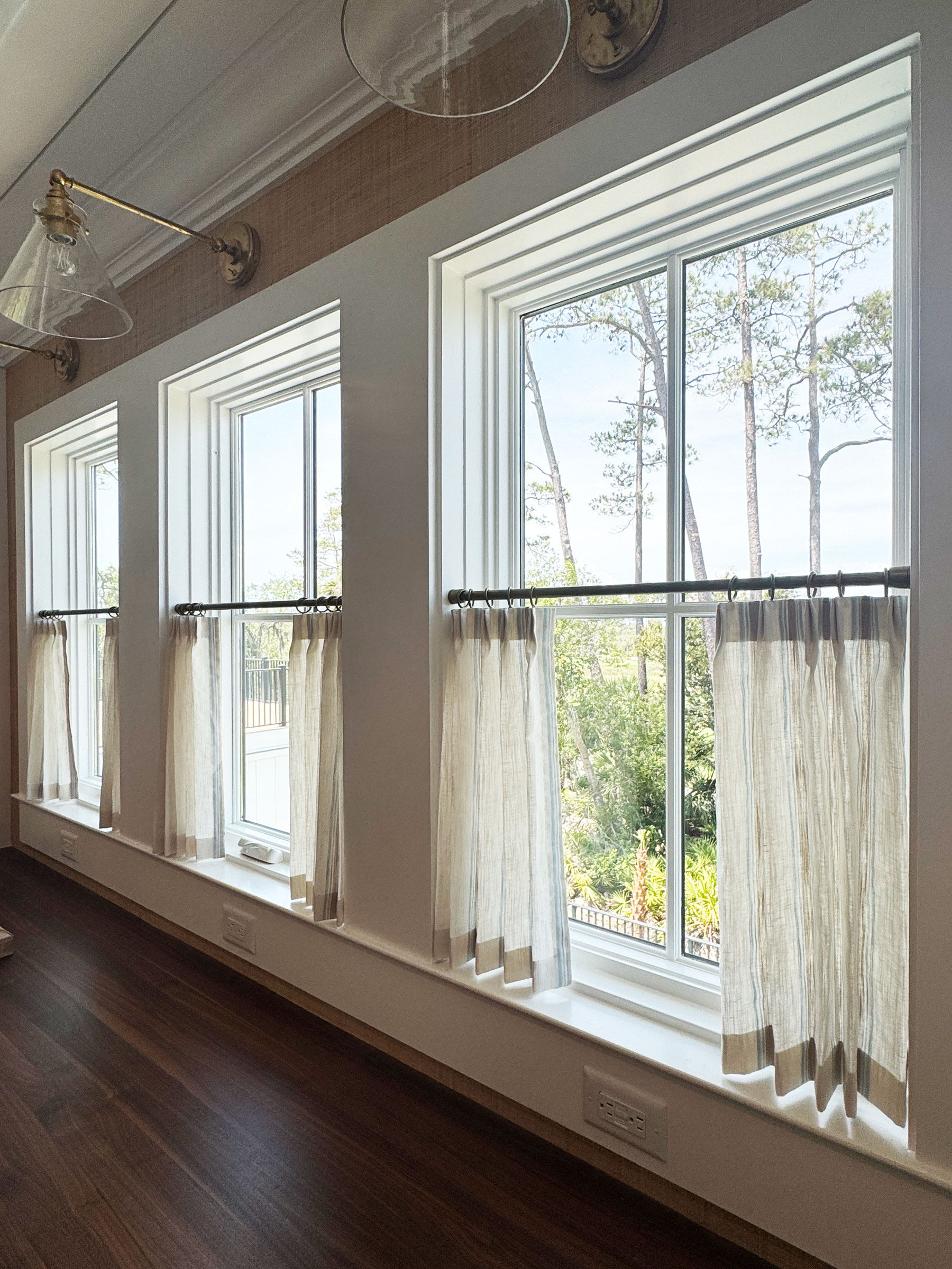 Three tall windows with white trim and half-length beige custom window treatments allow sunlight to enter a room with dark wooden floors. Trees and greenery are visible outside, hinting at a serene savannah landscape.