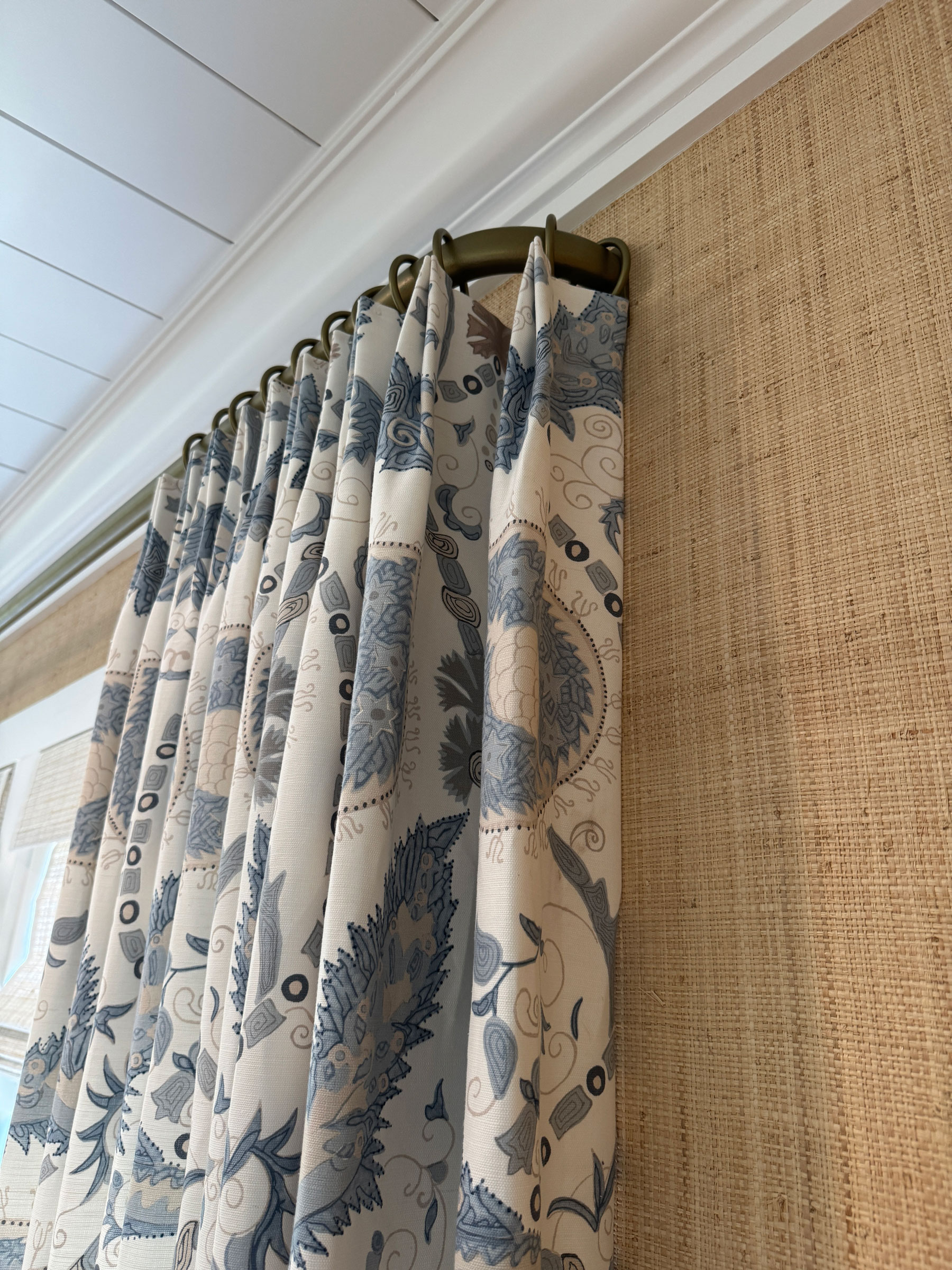 Blue and white patterned curtains, expertly selected window treatments, hang from a metal rod, set against a woven tan wall covering and crisp white ceiling trim, creating a refined design reminiscent of classic Savannah interiors.