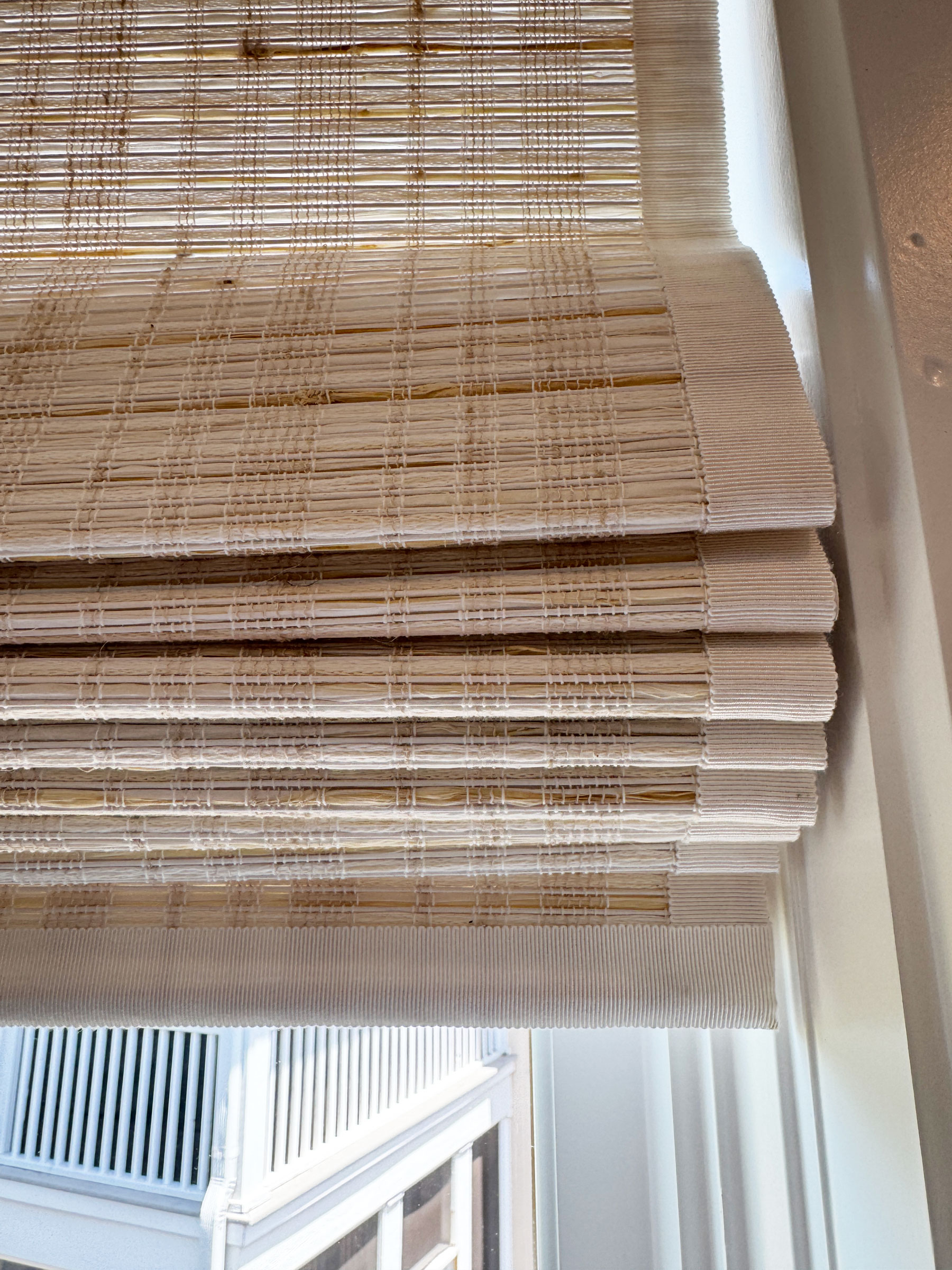 Close-up of a partially raised bamboo window shade with sunlight filtering through, revealing an exterior view of a building with white railings below—an inviting scene that highlights the harmonious design between natural materials and soft furnishings.