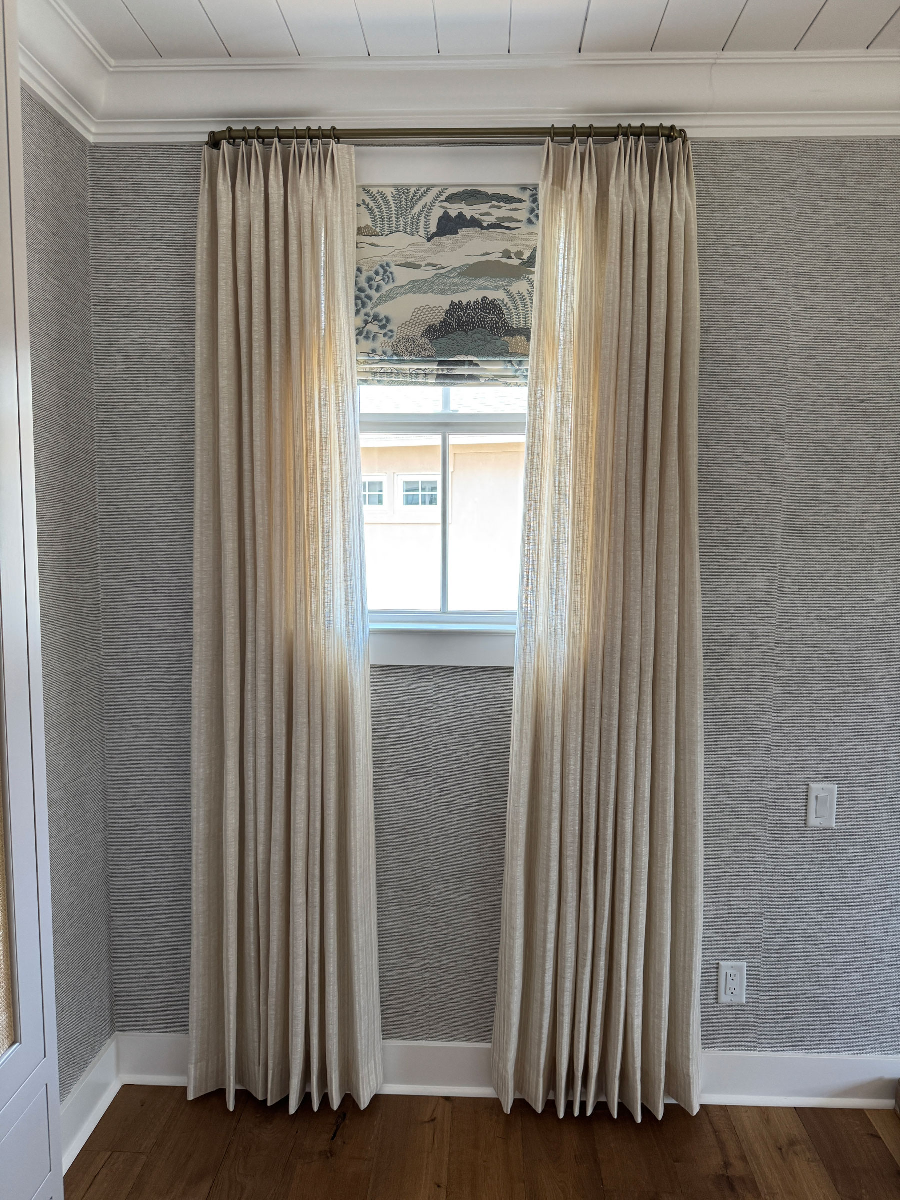 A window with cream-colored curtains and a decorative Roman shade featuring a landscape pattern, set in a room with gray textured walls and wood flooring, showcases elegant custom window treatments that beautifully complement the space’s soft furnishings.