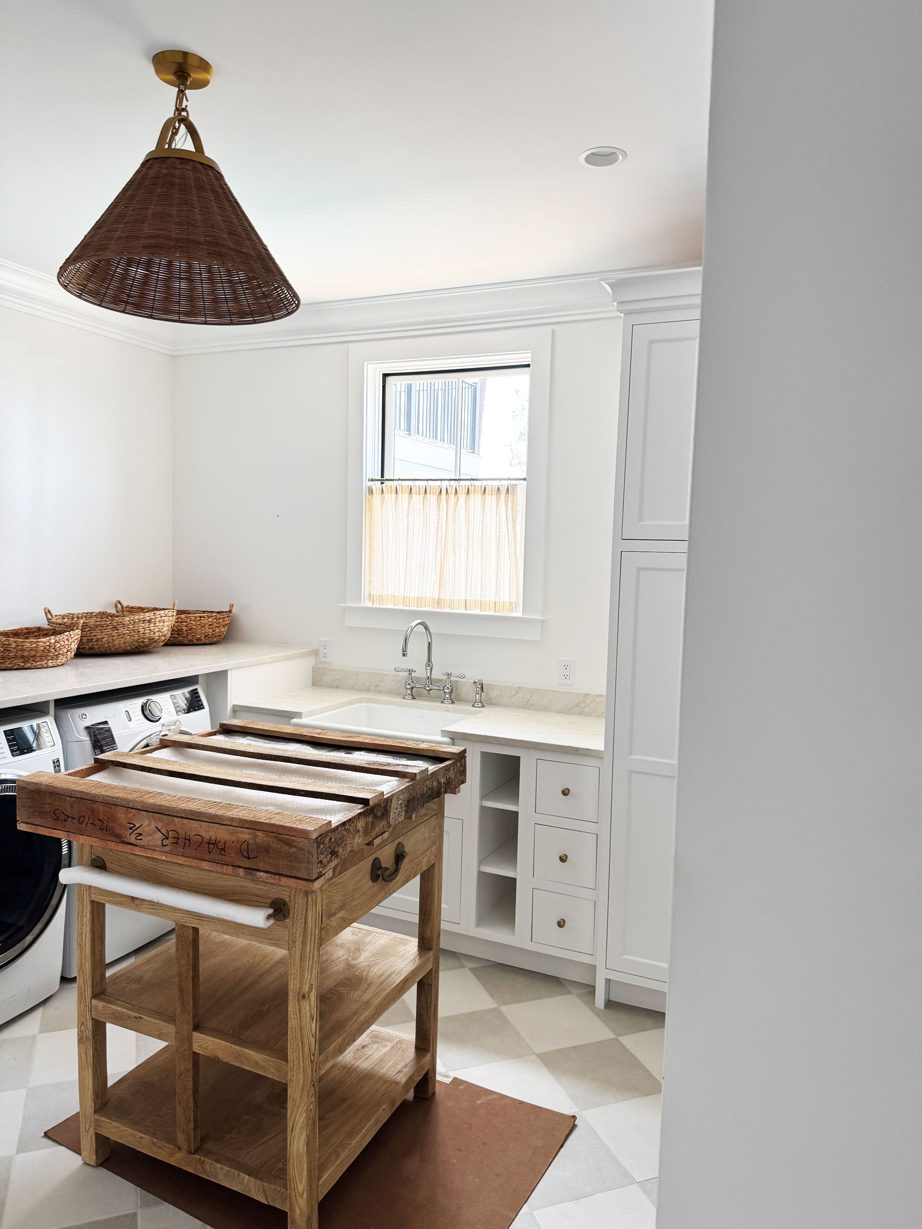 Laundry room with washer and dryer, a wooden cart in the center, wicker baskets on counter, sink below window with custom window treatments, and checkered floor tiles that complement the room’s inviting design.