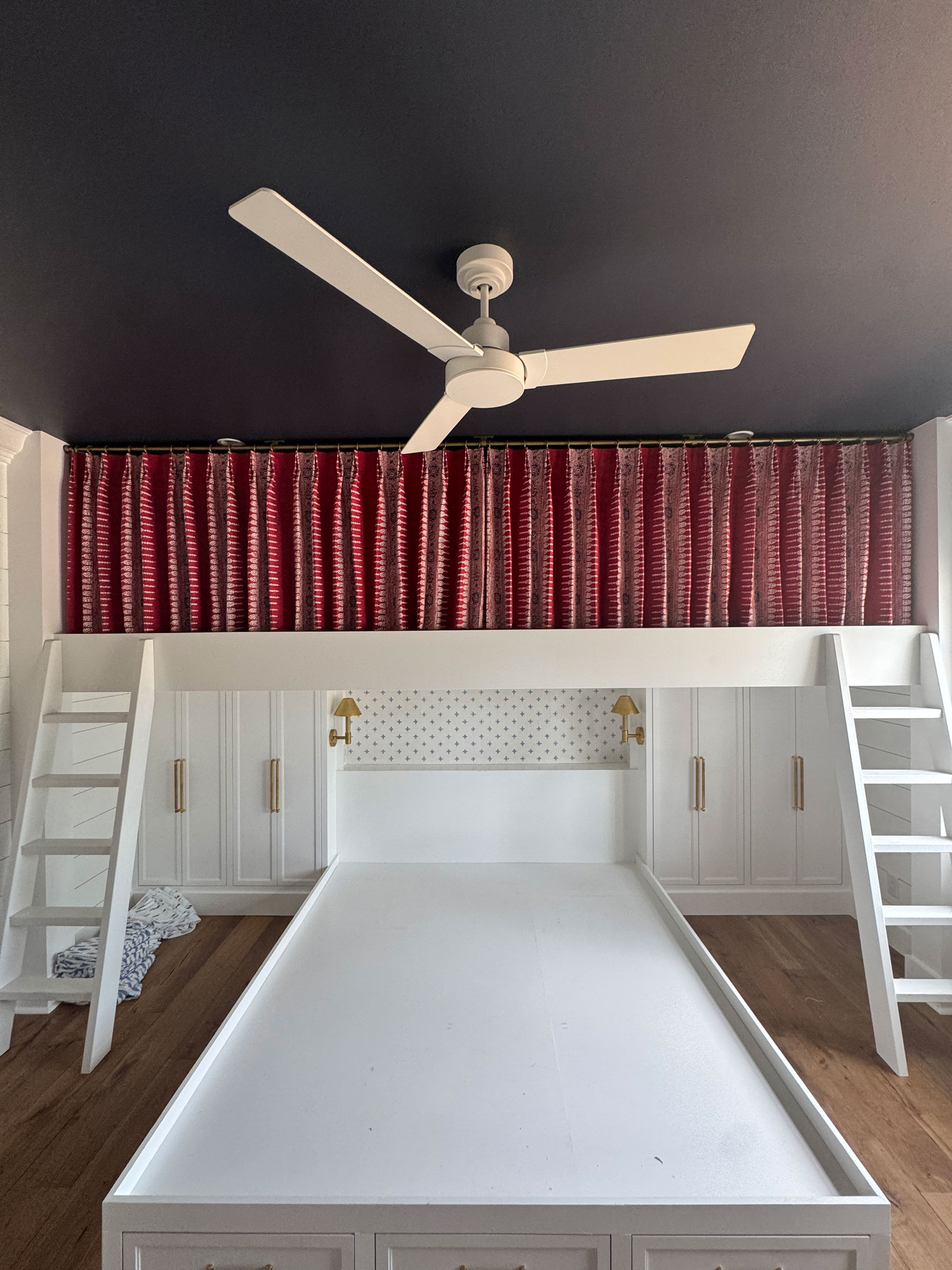 A white bunk bed with two ladders, a white ceiling fan, custom window treatments in red patterned curtains, and wood flooring in a bedroom.