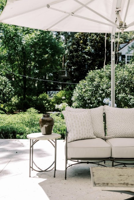 White cushioned outdoor sofa and small side table with a vase sit under a large umbrella on a patio in Savannah, with lush greenery, string lights in the background, and custom window treatments framing the nearby windows.